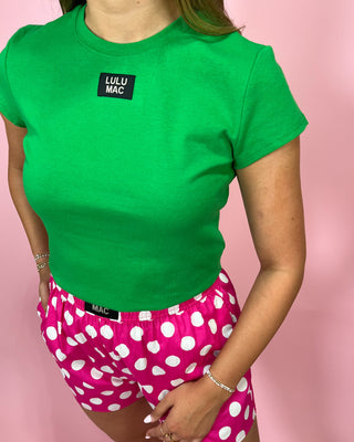 Person wearing a green t-shirt with 'LULU MAC' branding and pink polka dot shorts on a pink background.