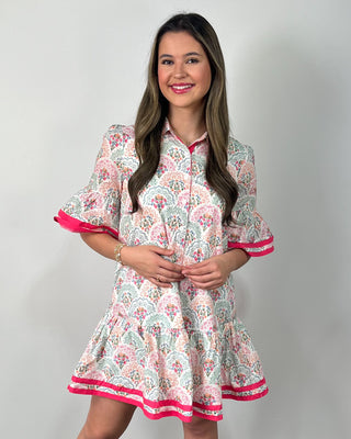 Smiling woman posing indoors wearing a playful printed swing dress with a collared neckline, button-front detail, and a flouncy tiered hem, featuring a soft floral-inspired pattern in pastel tones accented with bold hot pink trim on the ruffle sleeves and hem for the perfect pop, styled with clear slide sandals and simple gold jewelry for an easy, cheerful look.