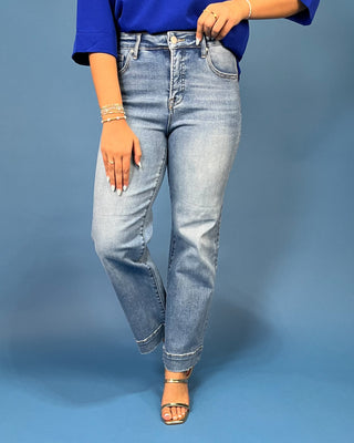 A person wearing light-wash blue denim jeans with a straight-to-relaxed leg, styled with a bright blue top and metallic sandals against a solid blue studio background.