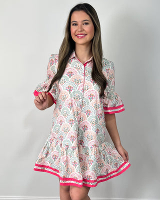 Smiling woman posing indoors wearing a playful printed swing dress with a collared neckline, button-front detail, and a flouncy tiered hem, featuring a soft floral-inspired pattern in pastel tones accented with bold hot pink trim on the ruffle sleeves and hem for the perfect pop, styled with clear slide sandals and simple gold jewelry for an easy, cheerful look.