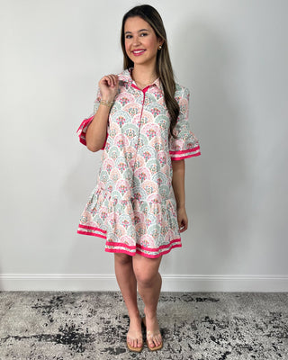 Smiling woman posing indoors wearing a playful printed swing dress with a collared neckline, button-front detail, and a flouncy tiered hem, featuring a soft floral-inspired pattern in pastel tones accented with bold hot pink trim on the ruffle sleeves and hem for the perfect pop, styled with clear slide sandals and simple gold jewelry for an easy, cheerful look.