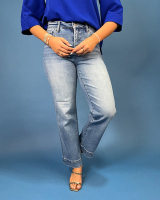 A person wearing light-wash blue jeans with a slight flare at the hem, styled with a bright blue top and metallic sandals, standing against a blue studio backdrop.