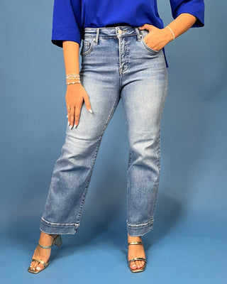 A model wearing light-wash blue denim jeans with a straight-to-medium leg, styled with a bright blue top and metallic sandals against a solid blue studio background.