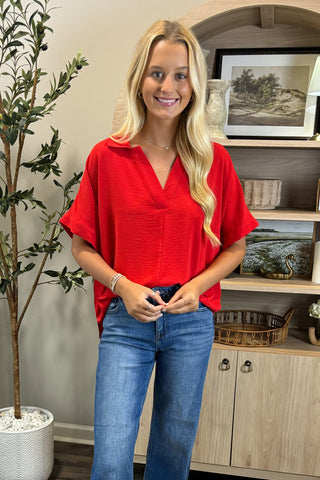 Woman wearing a red blouse and blue jeans standing in a room with a plant and shelves.