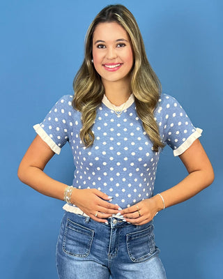  a light blue short-sleeve top with white polka dots and white ruffled trim, paired with blue jeans, standing against a solid blue background.