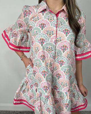 Smiling woman posing indoors wearing a playful printed swing dress with a collared neckline, button-front detail, and a flouncy tiered hem, featuring a soft floral-inspired pattern in pastel tones accented with bold hot pink trim on the ruffle sleeves and hem for the perfect pop, styled with clear slide sandals and simple gold jewelry for an easy, cheerful look.