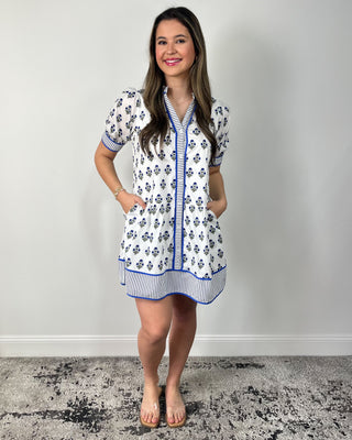 Smiling woman posing in front of a neutral backdrop wearing a white mini dress with a blue floral block print, puff sleeves, and a relaxed tiered silhouette, finished with striped blue trim along the neckline, sleeves, and hem, plus clear slide sandals and simple gold jewelry
