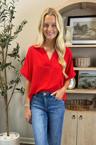 Woman wearing a red blouse and blue jeans standing indoors with decorative elements in the background.