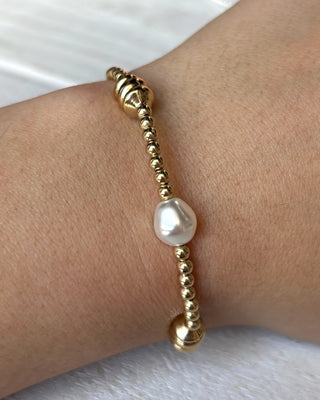 Gold beaded bracelet with a pearl on a wrist against a pink background