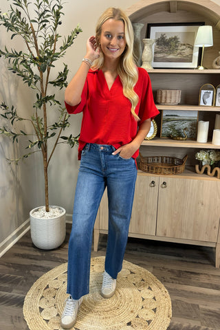 Woman in a red blouse and blue jeans standing in a room with a plant and wooden cabinet.