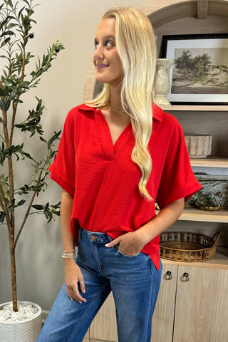 Woman wearing a red shirt and blue jeans standing indoors with decorative elements in the background.