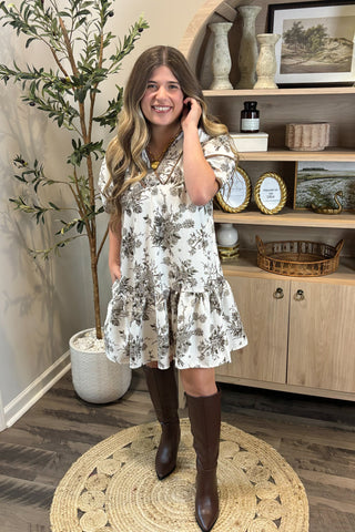 Woman in a floral dress and brown knee-high boots standing in a room with a plant and wooden cabinet.