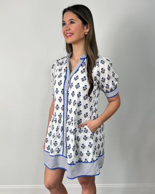 Smiling woman posing in front of a neutral backdrop wearing a white mini dress with a blue floral block print, puff sleeves, and a relaxed tiered silhouette, finished with striped blue trim along the neckline, sleeves, and hem, plus clear slide sandals and simple gold jewelry