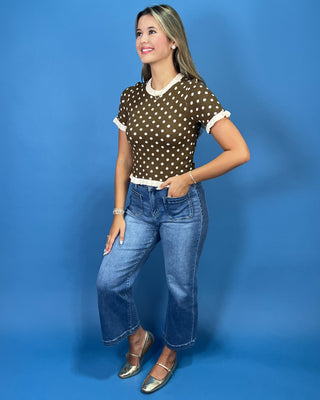 A woman modeling a brown short-sleeve top with white polka dots and white ruffled trim, paired with blue jeans and metallic silver shoes, against a solid blue studio background.