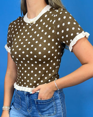 A woman wearing a brown short-sleeve knit top with white polka dots and white ruffled trim at the collar, sleeve cuffs, and hem, paired with blue jeans against a solid blue background.