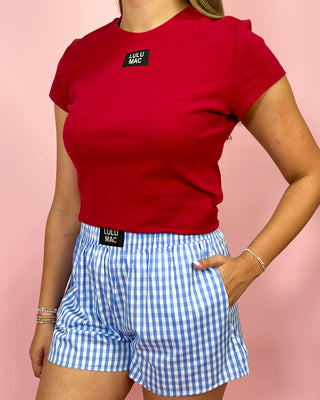 Person wearing a red t-shirt and blue checkered shorts with visible branding on a pink background