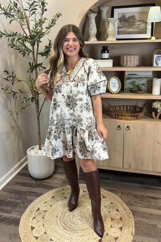 Woman in a floral dress and brown boots standing in a room with a wooden cabinet and decorative items.