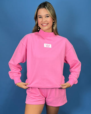 A woman modeling a bright pink sweatshirt and matching shorts set against a solid blue studio background. The sweatshirt features a mock neck and a small white rectangular chest patch with pink text