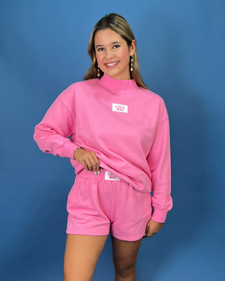 A woman modeling a bright pink two-piece lounge set consisting of a mock-neck sweatshirt and matching shorts, set against a solid blue studio background.