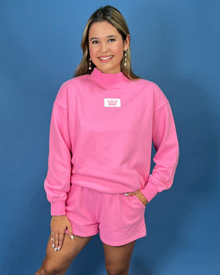 A woman modeling a bright pink mock-neck sweatshirt and matching shorts he sweatshirt features a small white rectangular chest patch
