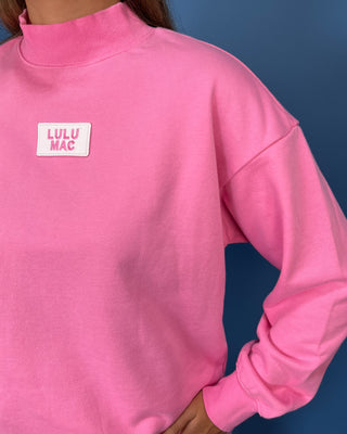 A close-up of a person wearing a bright pink mock-neck sweatshirt with a white rectangular patch on the left chest that reads 'LULU MAC' in pink letters, set against a dark blue background.