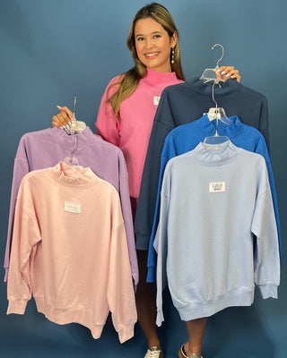 holding up four sweatshirts on hangers in different colors: lavender, light pink, navy, and light blue. Each sweatshirt has a small white rectangular chest label.