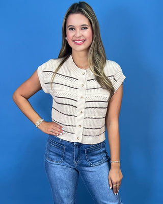 A woman modeling a cream-colored short-sleeve knit cardigan with thin brown horizontal stripes and button details, paired with blue jeans, set against a solid blue studio background.