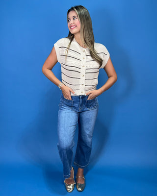 A woman posing in a cream-colored knit short-sleeve top with black horizontal stripes and blue jeans, standing against a solid blue studio background.