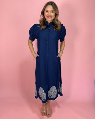 Woman wearing a navy dress with white embroidery on a pink background