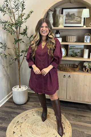 Woman in a burgundy dress standing in a room with a plant and shelves.