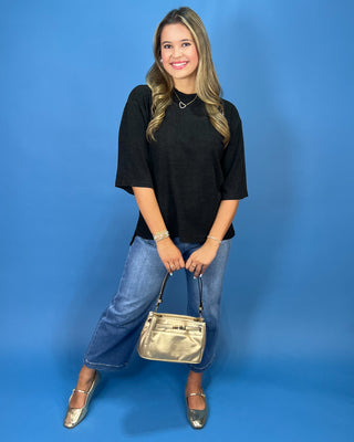 A woman modeling a black short-sleeve top, blue cropped jeans, and metallic gold flats, holding a metallic gold handbag against a solid blue studio backdrop.