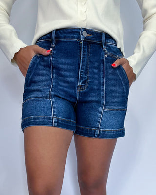 Dark wash denim shorts with front seam detail, cuffed hem, and high-rise fit paired with metallic sneakers.