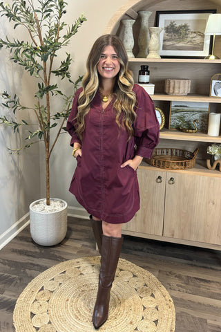 Woman in a burgundy dress and brown boots standing in a room with a plant and shelves.