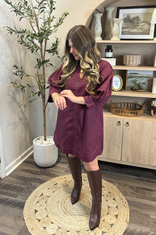 Woman in a purple dress and brown boots standing in a room with a plant and wooden cabinet.