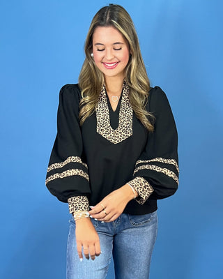 A woman wearing a black long-sleeve blouse with leopard print trim on the collar and cuffs, paired with blue jeans, standing against a solid blue background.