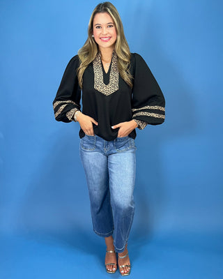 A woman modeling a black long-sleeve blouse with a leopard-print V-neckline and matching leopard-print sleeve cuffs, paired with blue jeans and metallic sandals, against a solid blue studio background.