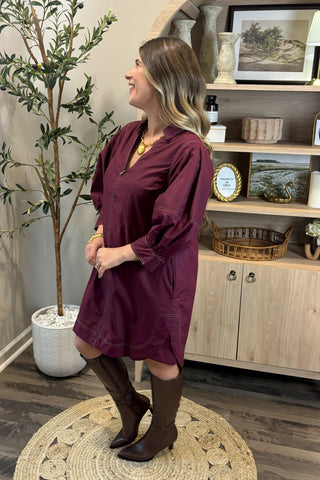 Woman in a burgundy dress standing in a room with a plant and wooden cabinet.
