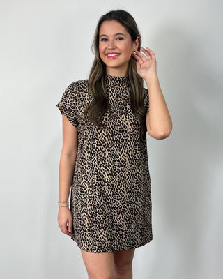 Smiling woman standing against a neutral background wearing a leopard print short-sleeve mini dress with a mock neckline, styled with clear slide sandals.