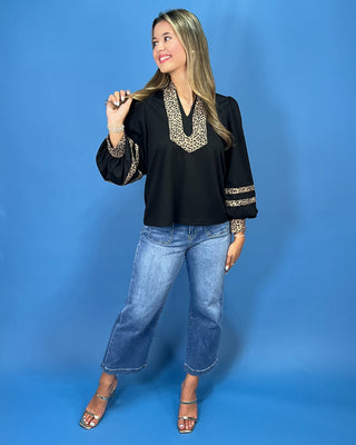 A woman modeling a black long-sleeve blouse with a leopard-print V-neck and leopard-print sleeve cuffs, paired with blue jeans and metallic sandals, against a solid blue studio background.