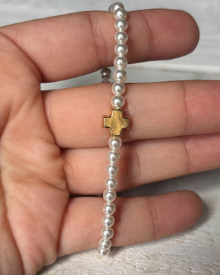 Pearl bracelet with a gold cross charm held in a hand against a white background