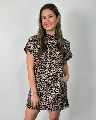 Smiling woman standing against a neutral background wearing a leopard print short-sleeve mini dress with a mock neckline, styled with clear slide sandals.