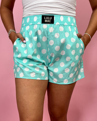 Person wearing teal shorts with white polka dots and a visible brand label on a pink background