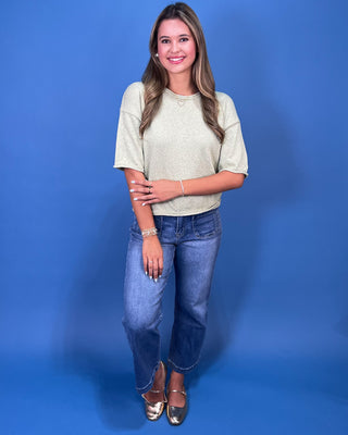 A woman modeling a light beige short-sleeve knit top and blue jeans against a solid blue studio background.