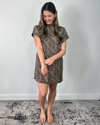 Smiling woman standing against a neutral background wearing a leopard print short-sleeve mini dress with a mock neckline, styled with clear slide sandals.