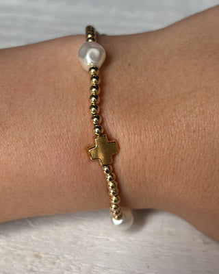 Gold beaded bracelet with a cross charm on a wrist against a neutral background