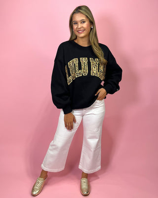Woman wearing a black sweatshirt with gold text, white pants, and gold shoes on a pink background