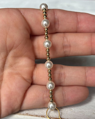 Gold and pearl beaded hair tie held between fingers on a neutral background