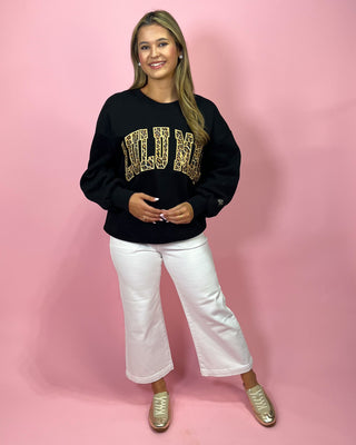 Woman wearing a black sweatshirt with gold lettering, white pants, and gold shoes on a pink background