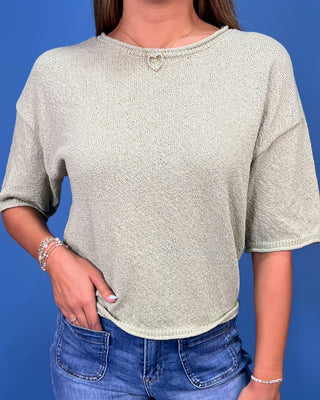 A woman wearing a light sage green knit short-sleeve crop top with a small silver heart detail at the neckline, paired with blue jeans, set against a solid blue background.
