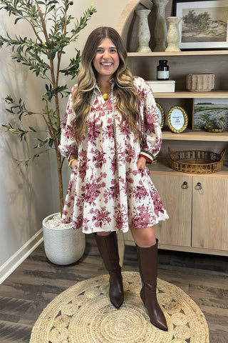 Woman in a floral dress standing in a room with wooden flooring and a plant.